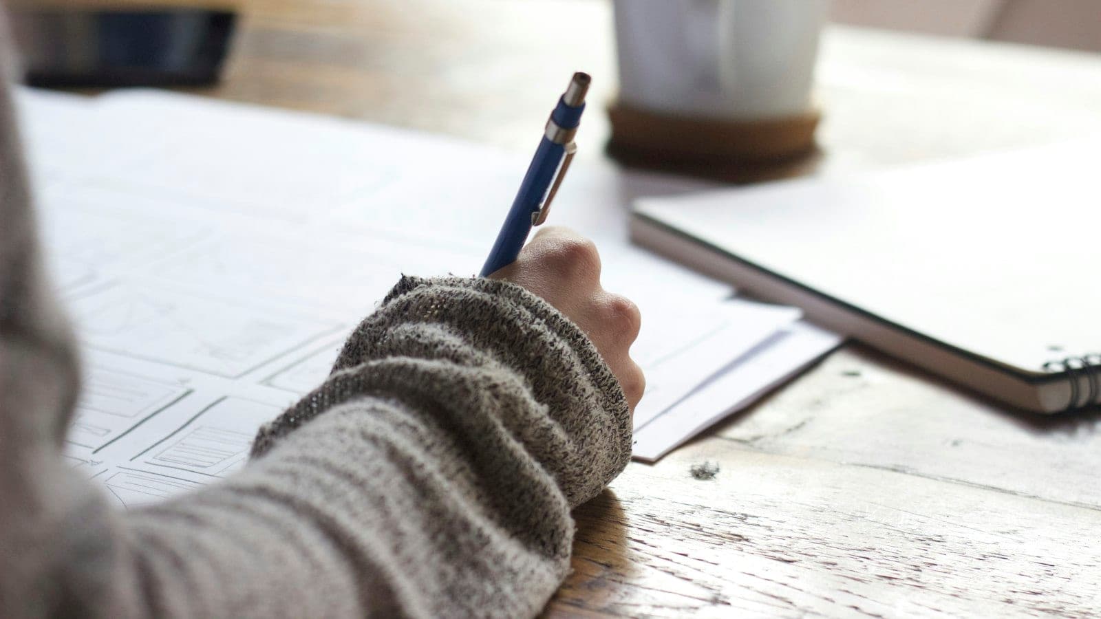 Close-up of a founder's hand sketching post ideas on index cards spread across a desk, with a phone and coffee cup nearby